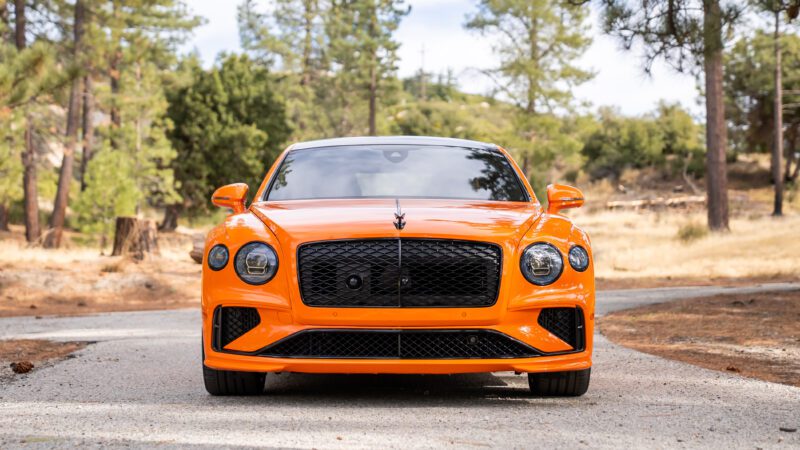 Front view of an orange Bentley car parked on a narrow outdoor path with trees and dry grass in the background.