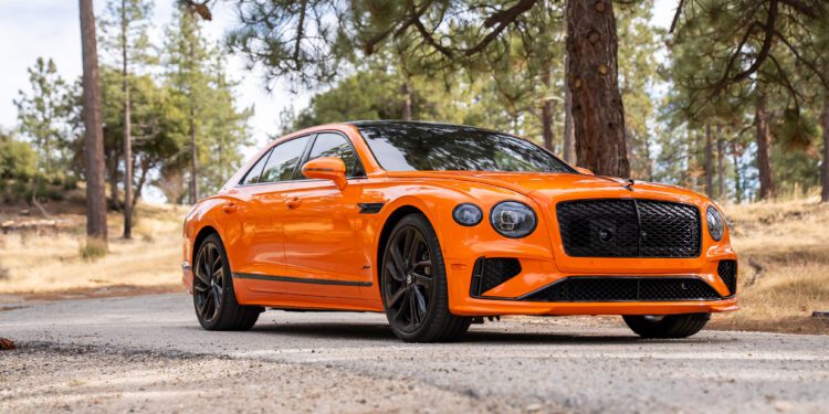 An orange luxury sedan with black accents parked on a forest road, surrounded by trees and dry grass.