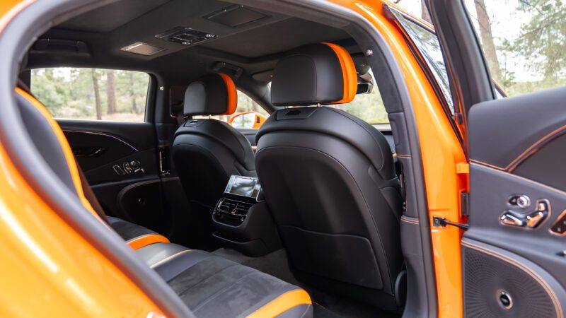 Rear view of a luxury car interior with black and orange leather seats, visible dashboard screens, and open rear door.
