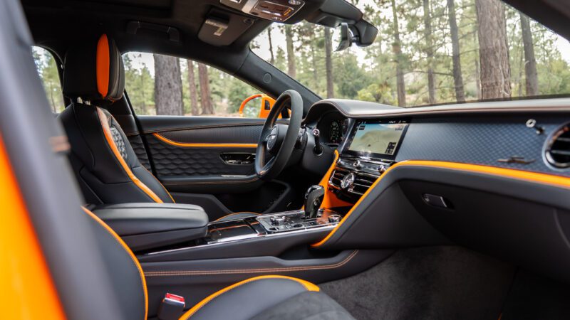 View of a luxury car interior featuring black leather seats with orange accents, a modern dashboard, touchscreen display, and trees visible through the windows.