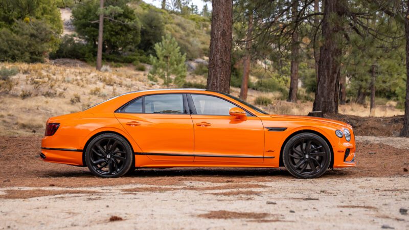 A bright orange luxury sedan with black wheels is parked outdoors among trees and dry grass.