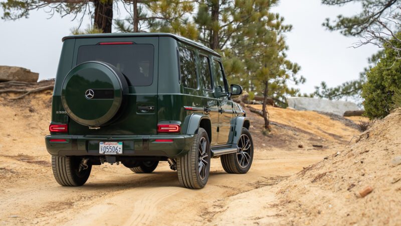 A green Mercedes-Benz G-Class SUV is parked on a dirt road surrounded by trees and rocks.