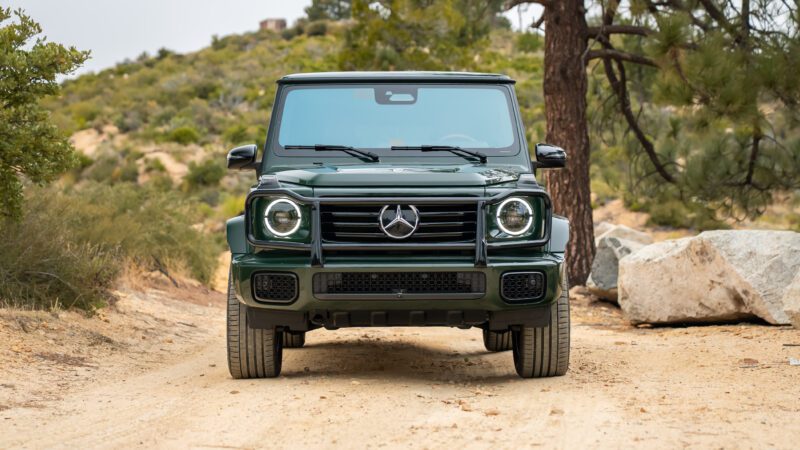 A front view of a green Mercedes-Benz G-Class SUV parked on a dirt road with trees and rocks in the background.