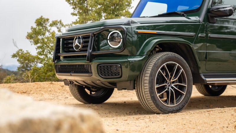 Front view of a green Mercedes-Benz G-Class SUV parked on a dirt surface, showing the grille, headlights, and front wheel. Trees are visible in the background.