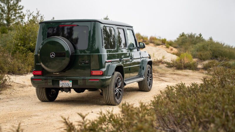 A dark green Mercedes-Benz G-Class SUV is parked on a dirt trail surrounded by greenery and shrubs.