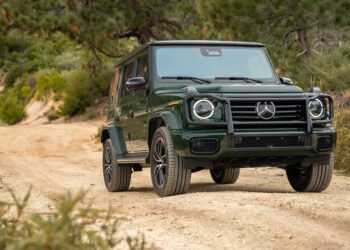 A green Mercedes-Benz G-Class SUV is parked on a dirt trail surrounded by trees and shrubs in a natural outdoor setting.