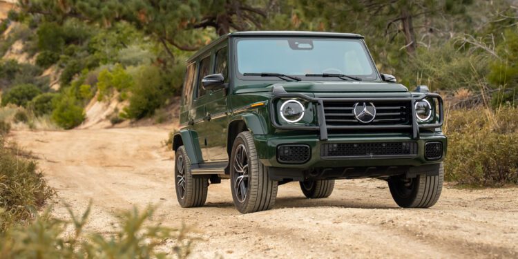 A green Mercedes-Benz G-Class SUV is parked on a dirt trail surrounded by trees and shrubs in a natural outdoor setting.
