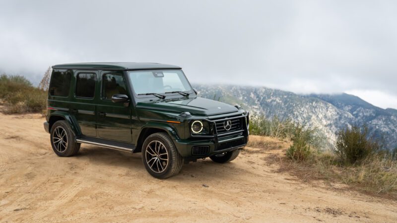 A dark green Mercedes-Benz G-Class SUV is parked on a dirt road with mountains and cloudy sky in the background.