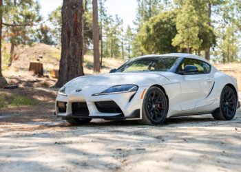 A white Toyota Supra sports car is parked on a dirt road surrounded by trees and natural landscape.