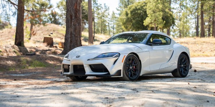 A white Toyota Supra sports car is parked on a dirt road surrounded by trees and natural landscape.
