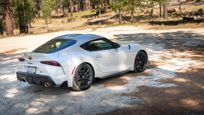 A silver Toyota Supra sports car is parked on a dirt clearing in a wooded area with trees and scattered sunlight.