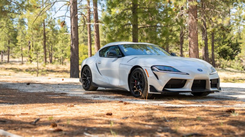 A white sports car is parked on a sunlit forest road surrounded by tall trees and scattered pine needles.
