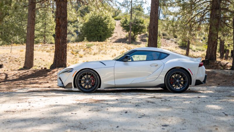 A silver sports car is parked on a dirt road in a wooded area with trees and bushes in the background.