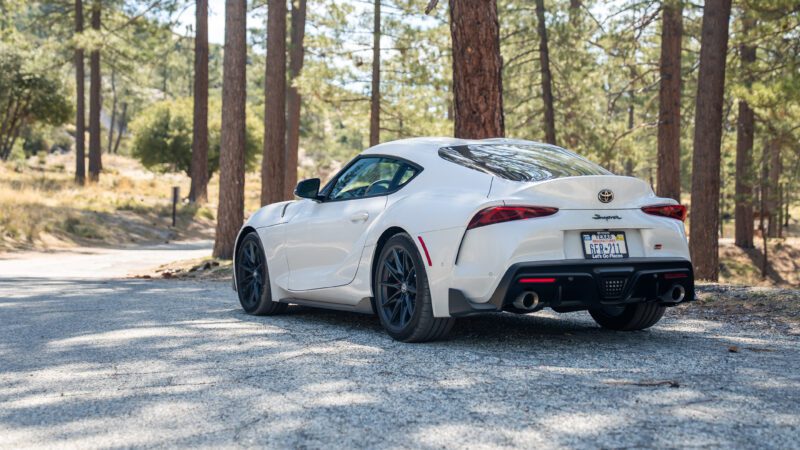 A white Toyota Supra sports car is parked on a gravel road in a forested area with tall trees and sunlight filtering through.