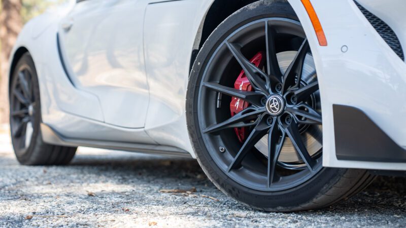 Close-up of a black alloy wheel with a red brake caliper on a white sports car parked on a gravel surface.