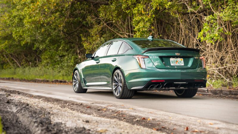 Green Cadillac sedan parked on a paved road next to trees, viewed from the rear left side.
