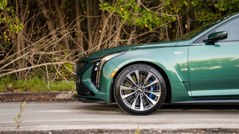 A green sports car parked on a road, with emphasis on the front wheel and headlight; trees and branches in the background.