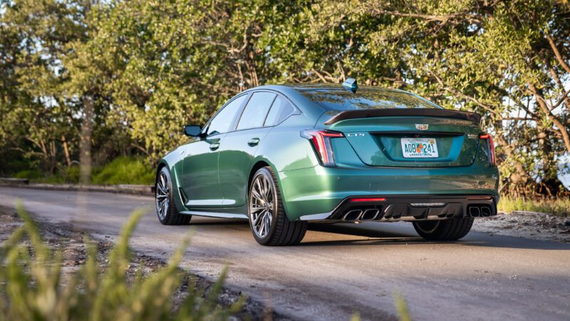 A green Cadillac sedan is parked on a paved road near trees, viewed from the rear left side.