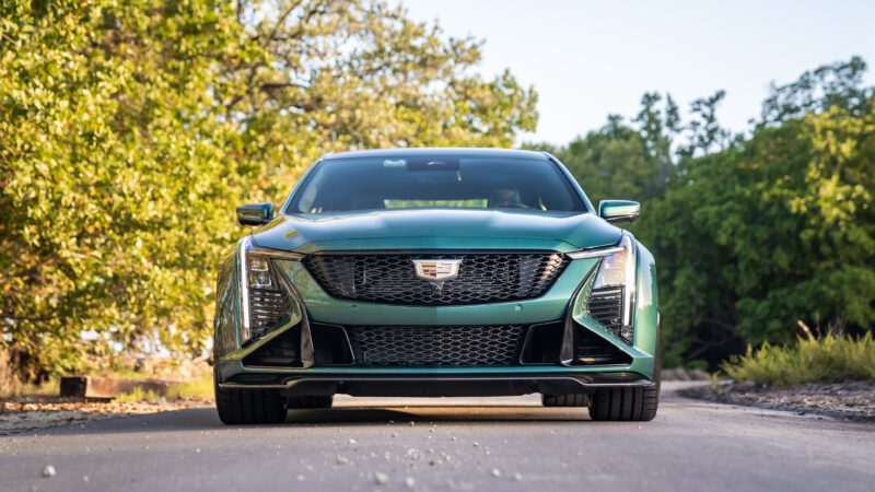 A front view of a green Cadillac sedan parked on a narrow road surrounded by trees on both sides.
