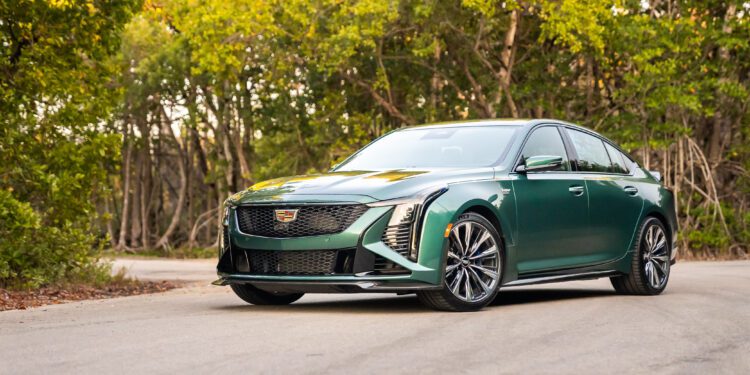 A green Cadillac sedan is parked on a paved road surrounded by trees and greenery.