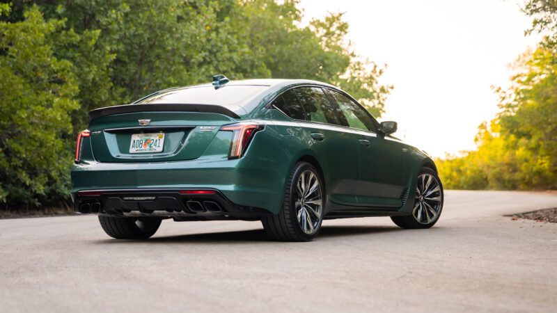 A green Cadillac sedan is parked on a road surrounded by trees, viewed from the rear left side.