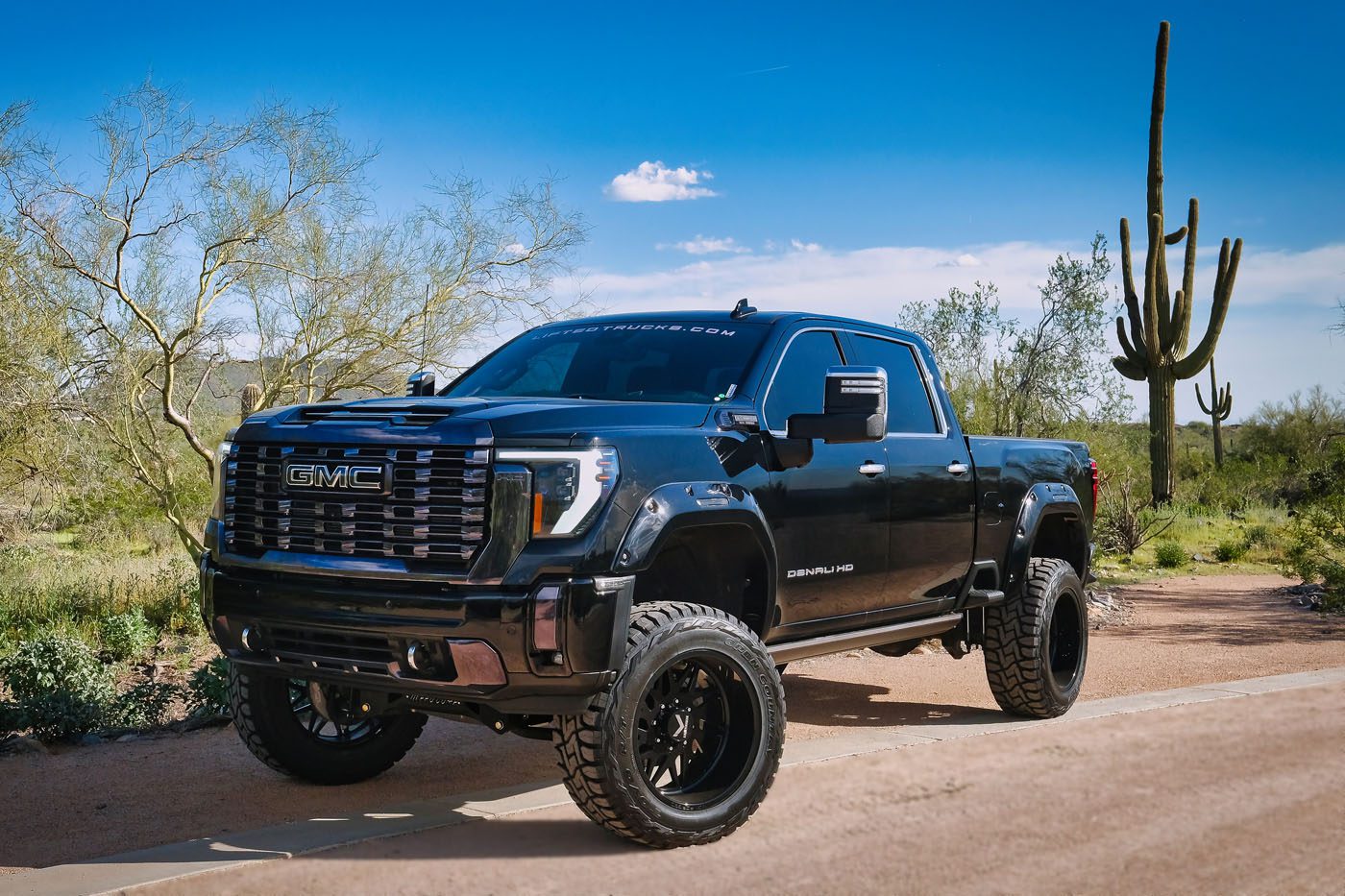 A black lifted GMC Sierra Denali pickup truck, from a top Lifted Truck Dealer, is parked on a paved road in a desert landscape with cacti and sparse vegetation under a clear blue sky.