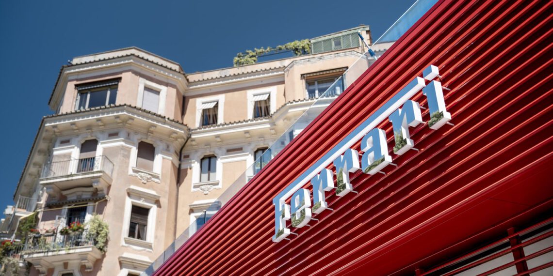 A red Ferrari storefront with sleek dealer corporate identity features a modern design in front of a beige, multi-story historic building under a clear blue sky.