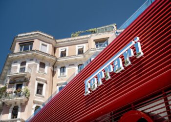 A red Ferrari storefront with sleek dealer corporate identity features a modern design in front of a beige, multi-story historic building under a clear blue sky.