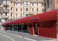 A modern red Bugatti showroom with horizontal slats and large glass windows, reflecting strong dealer corporate identity, stands on a city street lined with historic beige buildings.
