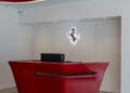 A modern reception desk in glossy red sits in front of a wall with a lit Ferrari logo, reflecting the dealer corporate identity in a minimalist, white office space.