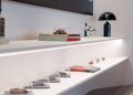 A row of Ferrari model cars in clear display cases is arranged on a white shelf beneath a large framed photo and several books, adding a touch of Dealer Corporate Identity to the modern, minimalist room.