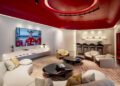 Modern lounge with cream chairs, round tables, a bar area, and a large TV showing a group in red sports uniforms; Ferrari-inspired red ceiling accents elevate the contemporary design and reflect sophisticated Dealer Corporate Identity.