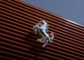 A silver prancing horse emblem, symbolizing Ferrari's corporate identity, is mounted on a horizontal red metal slatted wall, set against a clear blue sky.
