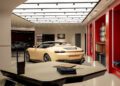 A beige Ferrari convertible sports car is displayed in a modern, well-lit showroom with red walls, automotive samples on shelves, and a distinct Dealer Corporate Identity throughout the space.