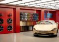 A gold Ferrari convertible sports car is displayed inside a showroom with red walls, showcasing dealer corporate identity, alongside paint samples, interior swatches, and wheels mounted on the wall.