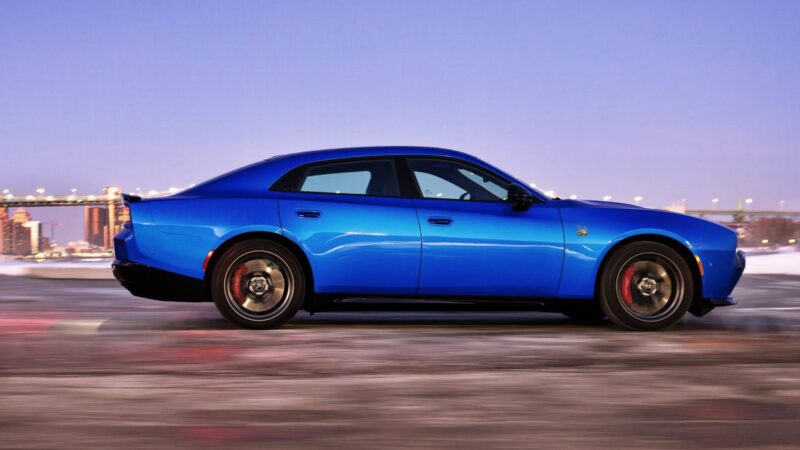 A blue four-door sports sedan drives on a road at dusk, city lights and a bridge in the background—reminding us the Four-Door Dodge Charger Daytona is here, but there’s more to this muscle car sedan than meets the eye.
