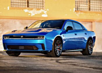 A blue modern sports sedan, inspired by the Four-Door Dodge Charger Daytona, with a sleek design is parked in front of a textured, yellow-painted brick wall—there's more to this muscle car sedan than meets the eye.