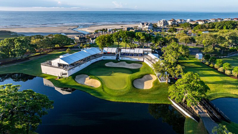 Aerial view of a coastal golf course with a green surrounded by sand bunkers, water hazards, grandstands, and nearby houses, with the ocean in the background.