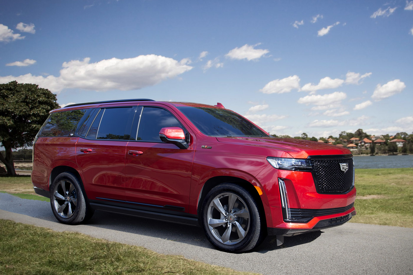A red SXR Series Cadillac SUV is parked on a paved path by the river, with trees, grass, and a partly cloudy sky in the background.