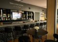 A modern bar area with black stools and a table with white flowers, featuring a large mirror behind the counter and "The Concours Club" sign on the wall.