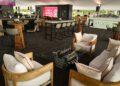 Modern lounge area with wooden chairs, glass table featuring a mechanical engine centerpiece, bar seating, high tables, and a prominent digital display in the background.