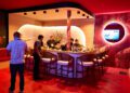 A group of people stand and sit around a bar counter in a modern, red-lit restaurant with patterned walls and a TV screen.