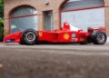 A red Ferrari Formula 1 car with sponsor logos is parked on a paved surface in front of a brick building, reminiscent of the Historic Monaco Sale where the Ferrari F2001 became the most valuable Schumacher-F1 car auctioned at €15.98 million.