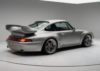A silver sports car with a large rear wing, considered one of the most expensive Porsches of all time, is parked in a white studio, viewed from the rear left side.