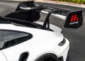 Close-up of a large rear wing with "Manthey" branding on a white sports car, parked on a paved surface with greenery in the background.