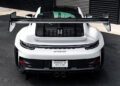 Rear view of a white Porsche 911 GT3 RS with a large rear wing, parked on a blacktop surface near a building with black and white panels.