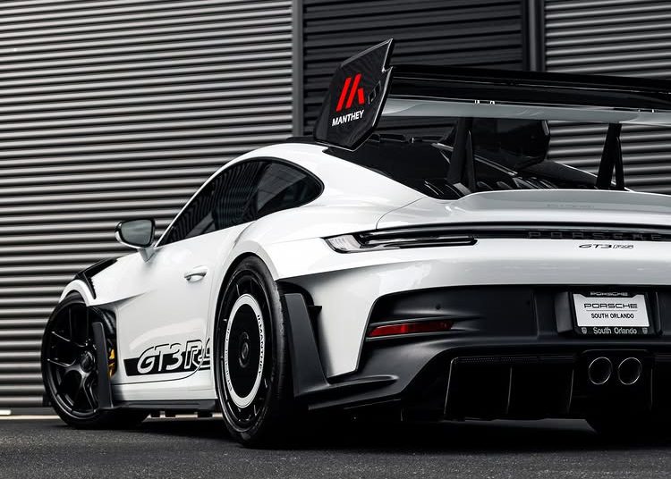 A white Porsche 911 GT3 RS with a large rear wing is parked in front of a building with metal siding.