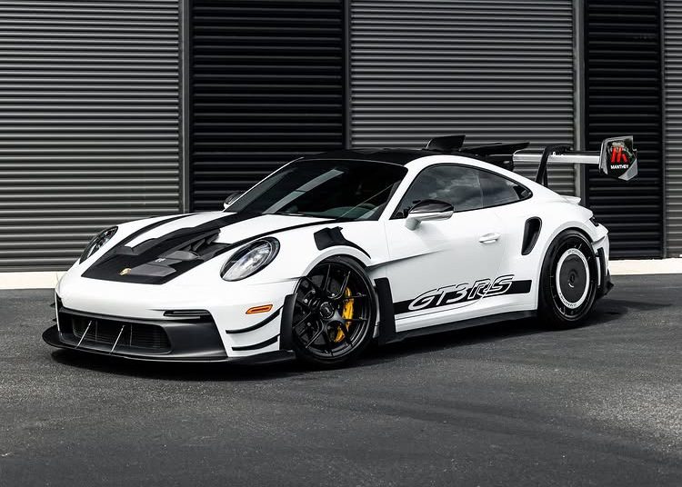 A white Porsche 911 GT3 RS with black accents and a large rear wing is parked in front of a modern building with dark horizontal panels.