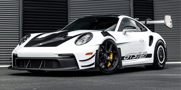 A white Porsche 911 GT3 RS sports car with black accents and a large rear wing is parked in front of a modern, striped metal wall.