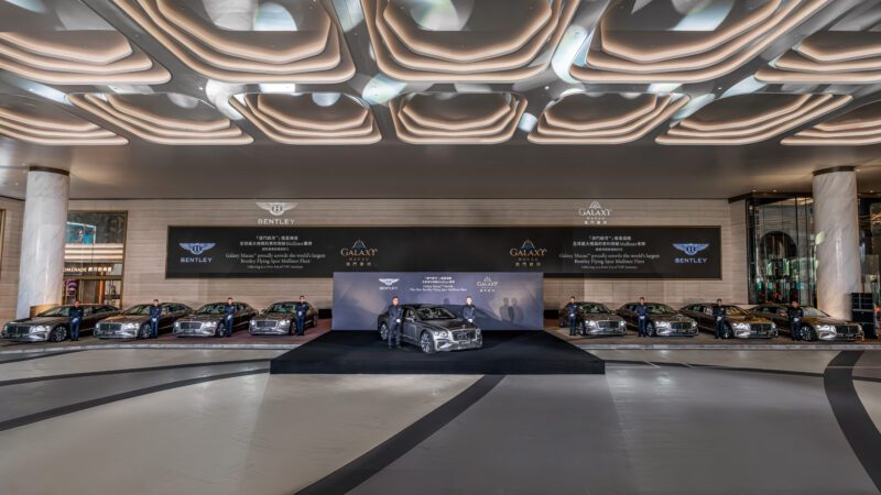 A row of Bentley Motors cars is displayed indoors on a showroom floor, with a Bespoke Flying Spur showcased on a central platform in front of a branded backdrop.